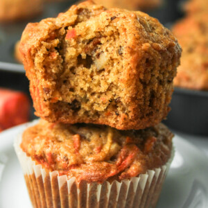 carrot apple muffin with a bite taken out of it stacked on top of another in a paper liner on a white ceramic plate