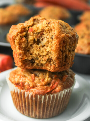 carrot apple muffin with a bite taken out of it stacked on top of another in a paper liner on a white ceramic plate
