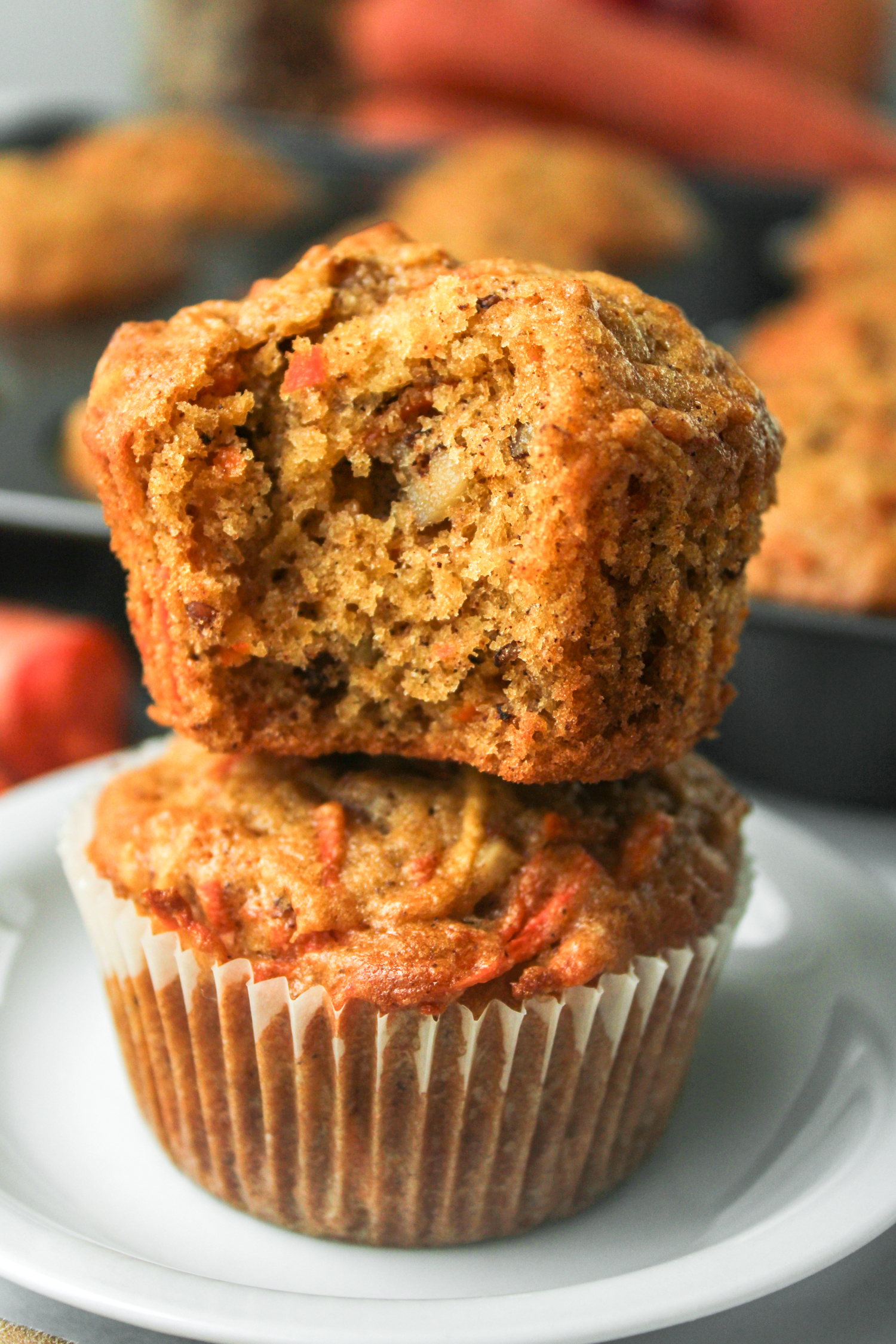 carrot apple muffin with a bite taken out of it stacked on top of another in a paper liner on a white ceramic plate