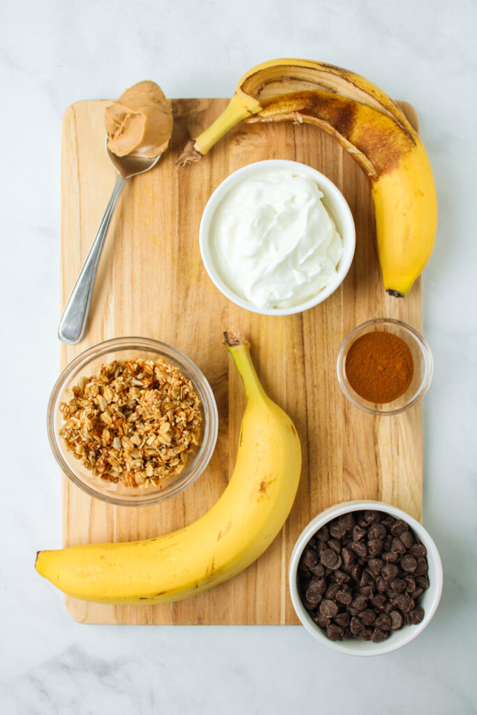 ingredients to a yogurt bowl on a wooden cutting board that includes peanut butter, banana, yogurt, granola, chocolate chips and cinnamon