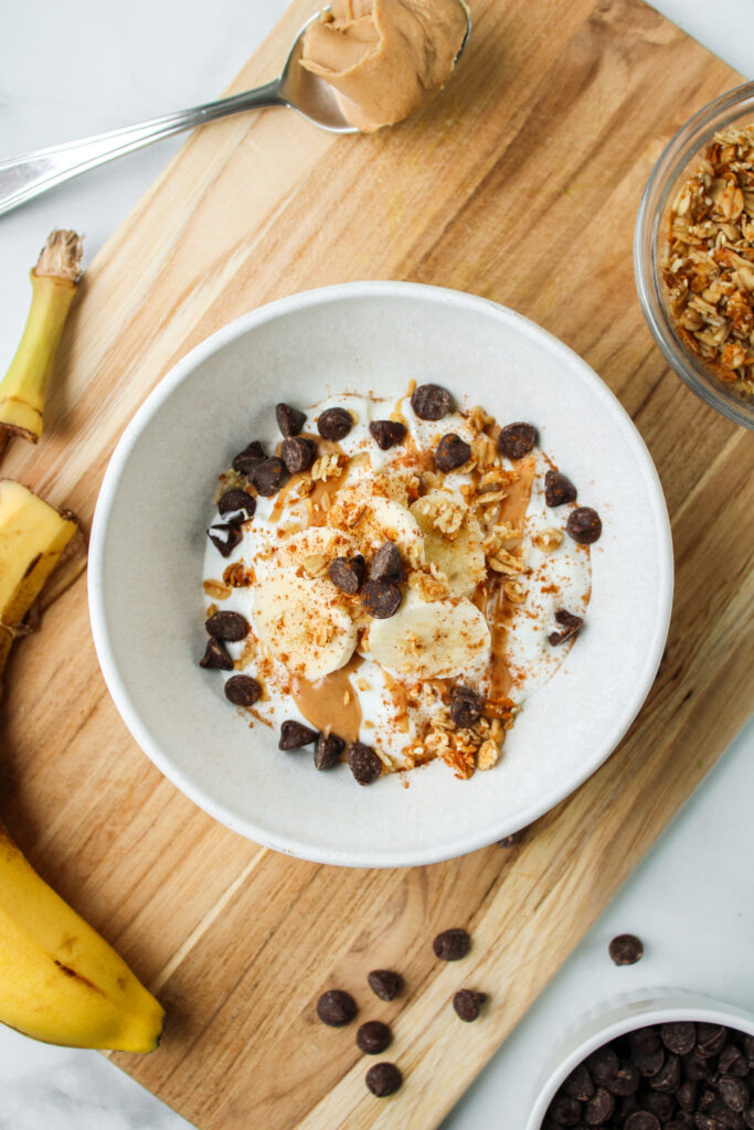 greek yogurt with peanut butter, banana, granola, chocolate chips and cinnamon in a white ceramic bowl on a wooden cutting board