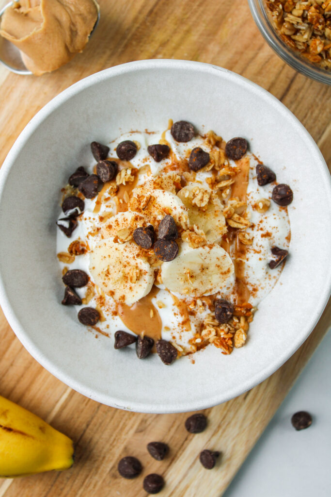 greek yogurt with peanut butter, banana, granola, chocolate chips and cinnamon in a white ceramic bowl on a wooden cutting board