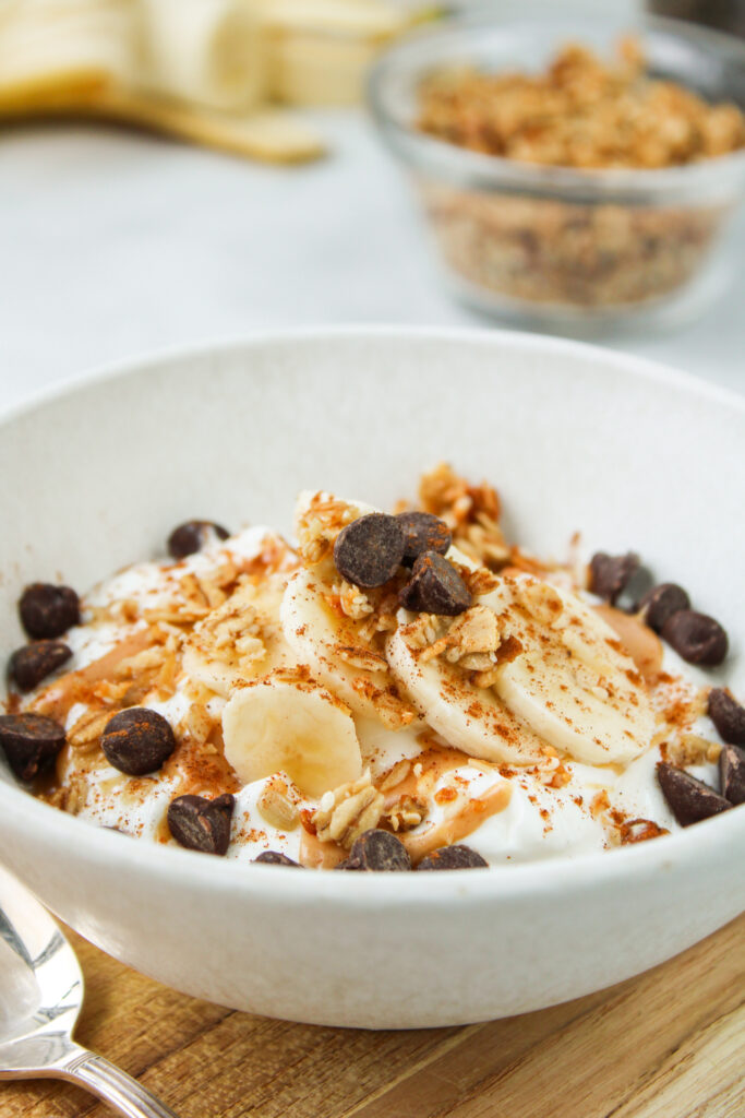 side view of greek yogurt with peanut butter, banana, granola, chocolate chips and cinnamon in a white ceramic bowl on a wooden cutting board