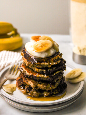 almond flour banana pancakes stacked on top of a white plate and topped with greek yogurt, banana and maple syrup