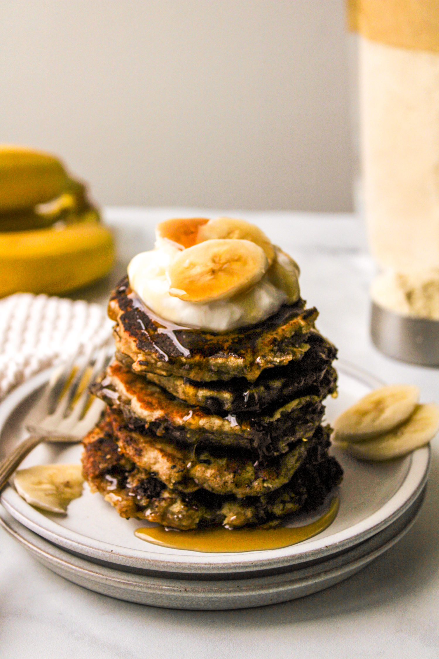 almond flour banana pancakes stacked on top of a white plate and topped with greek yogurt, banana and maple syrup