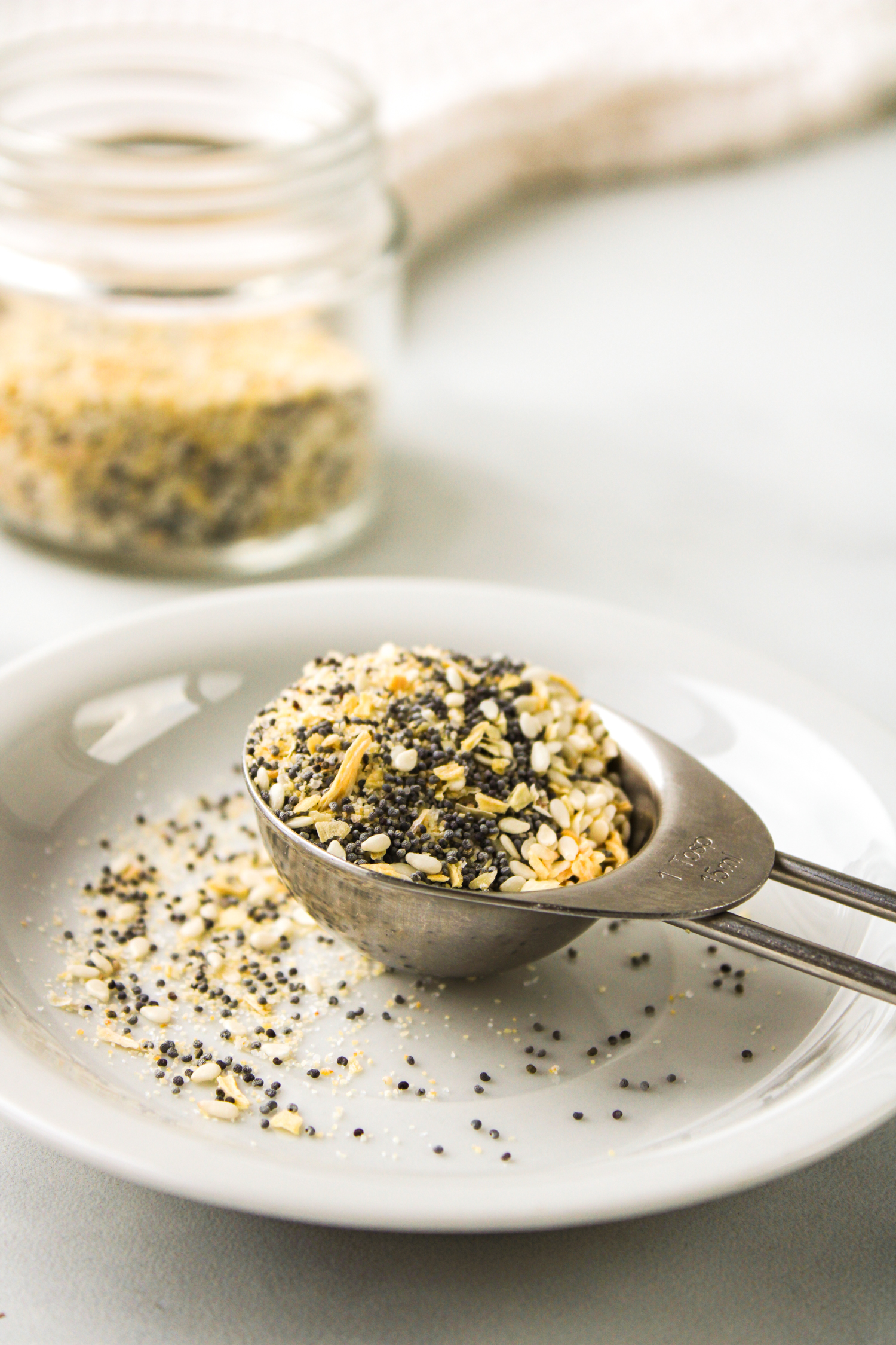profile shot of everything but the bagel seasoning in a metal measuring spoon on top of a small white plate