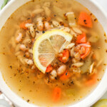 zoomed in overhead shot of vegan lemon orzo soup with a slice of lemon on top, in a white ceramic bowl with ingredients surrounding it