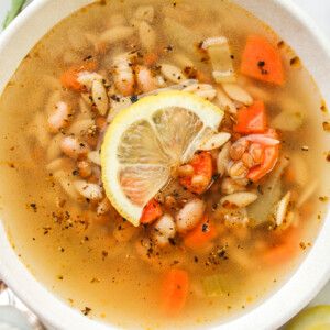 zoomed in overhead shot of vegan lemon orzo soup with a slice of lemon on top, in a white ceramic bowl with ingredients surrounding it