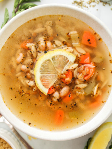 zoomed in overhead shot of vegan lemon orzo soup with a slice of lemon on top, in a white ceramic bowl with ingredients surrounding it