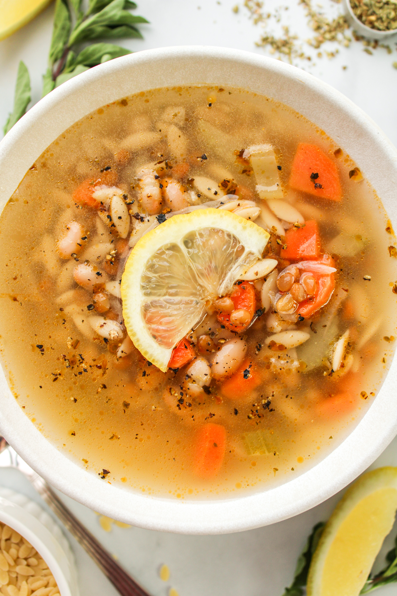 zoomed in overhead shot of vegan lemon orzo soup with a slice of lemon on top, in a white ceramic bowl with ingredients surrounding it