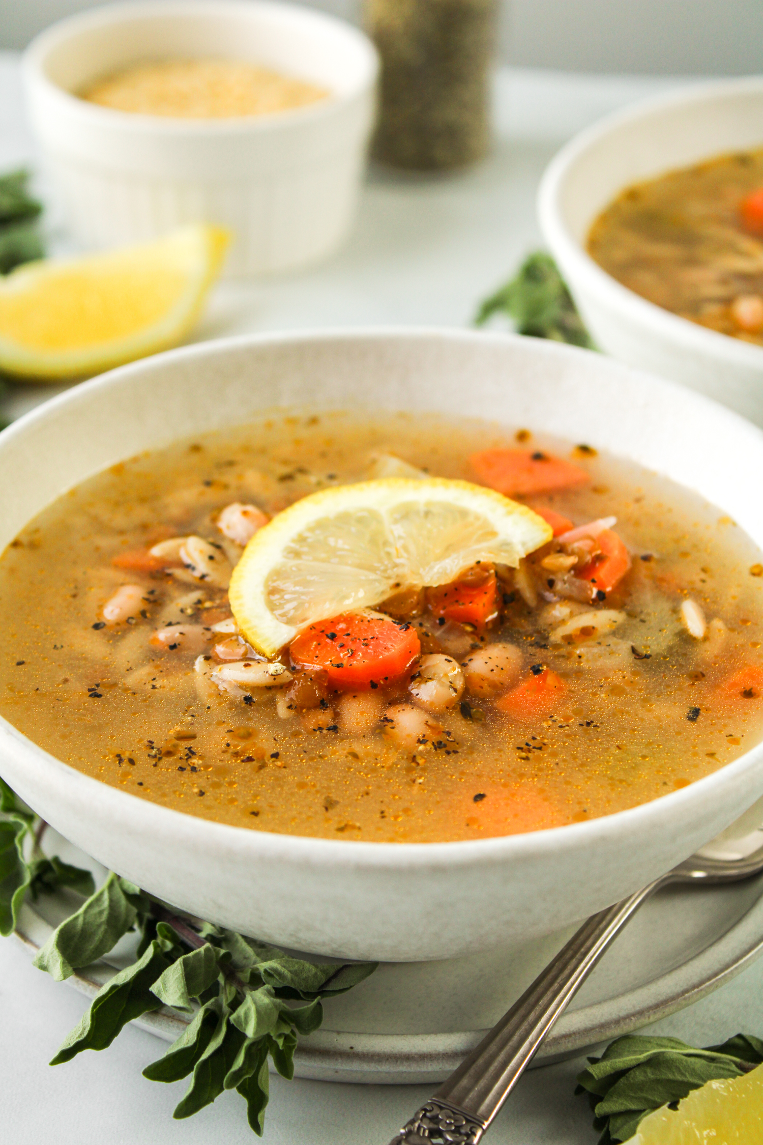 zoomed in side profile shot of lemon orzo soup with a slice of lemon on top, in a white ceramic bowl with ingredients in the background