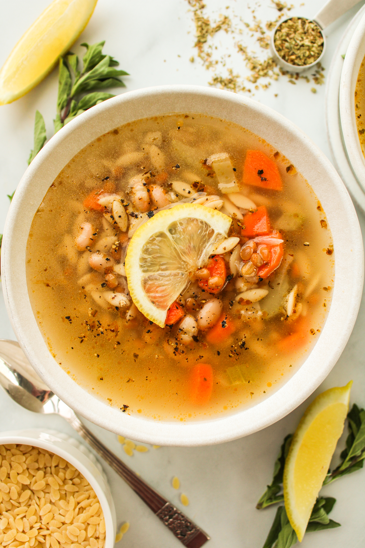 overhead shot of vegan lemon orzo soup with a slice of lemon on top, in a white ceramic bowl with ingredients surrounding it