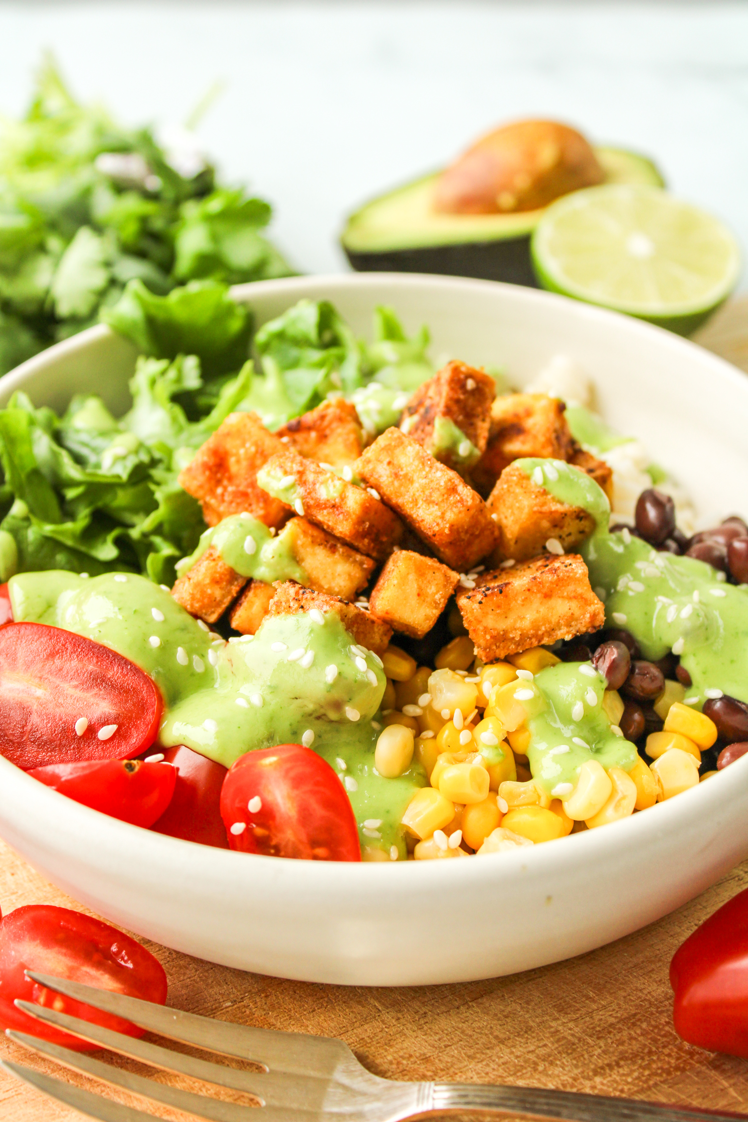side profile shot of tofu burrito bowl with avocado cilantro sauce drizzled on top in a white ceramic dish and avocado, lime and cilantro in the background