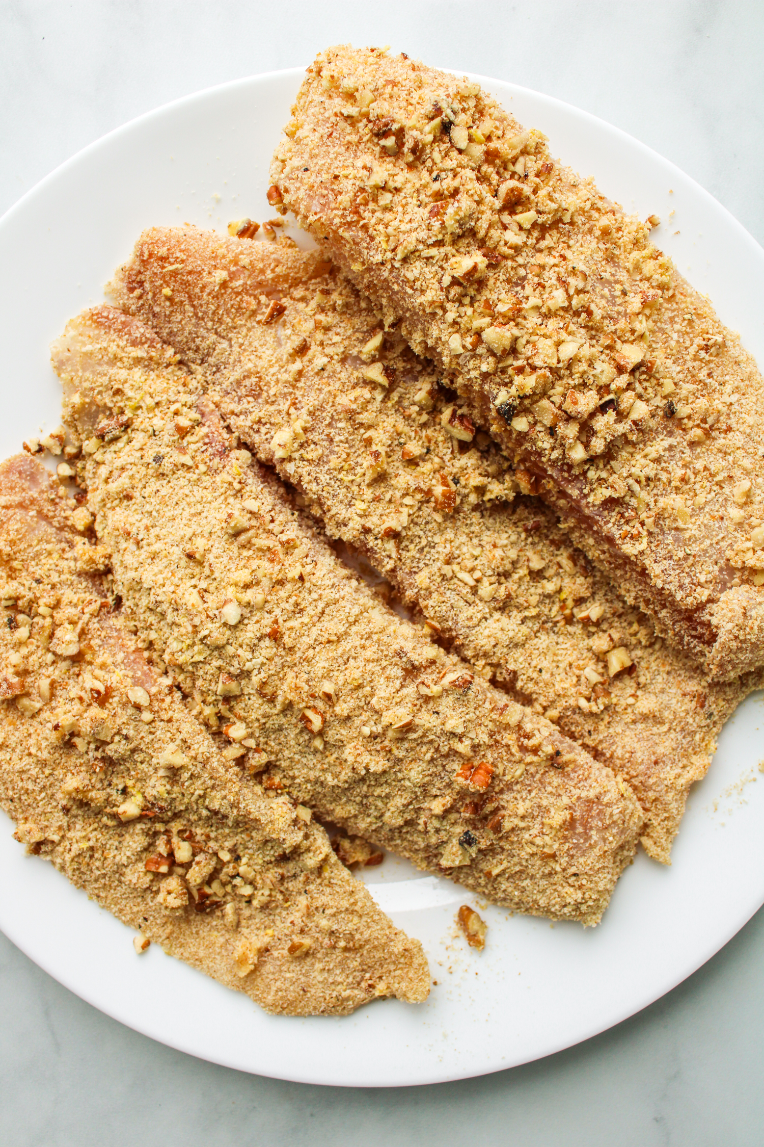 4 fresh fillets of breaded tilapia on a white plate before being cooked