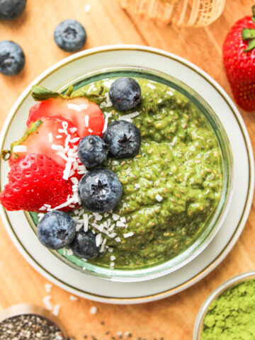 matcha chia seed pudding in a small glass bowl topped with berries and coconut flakes on top of a wooden board