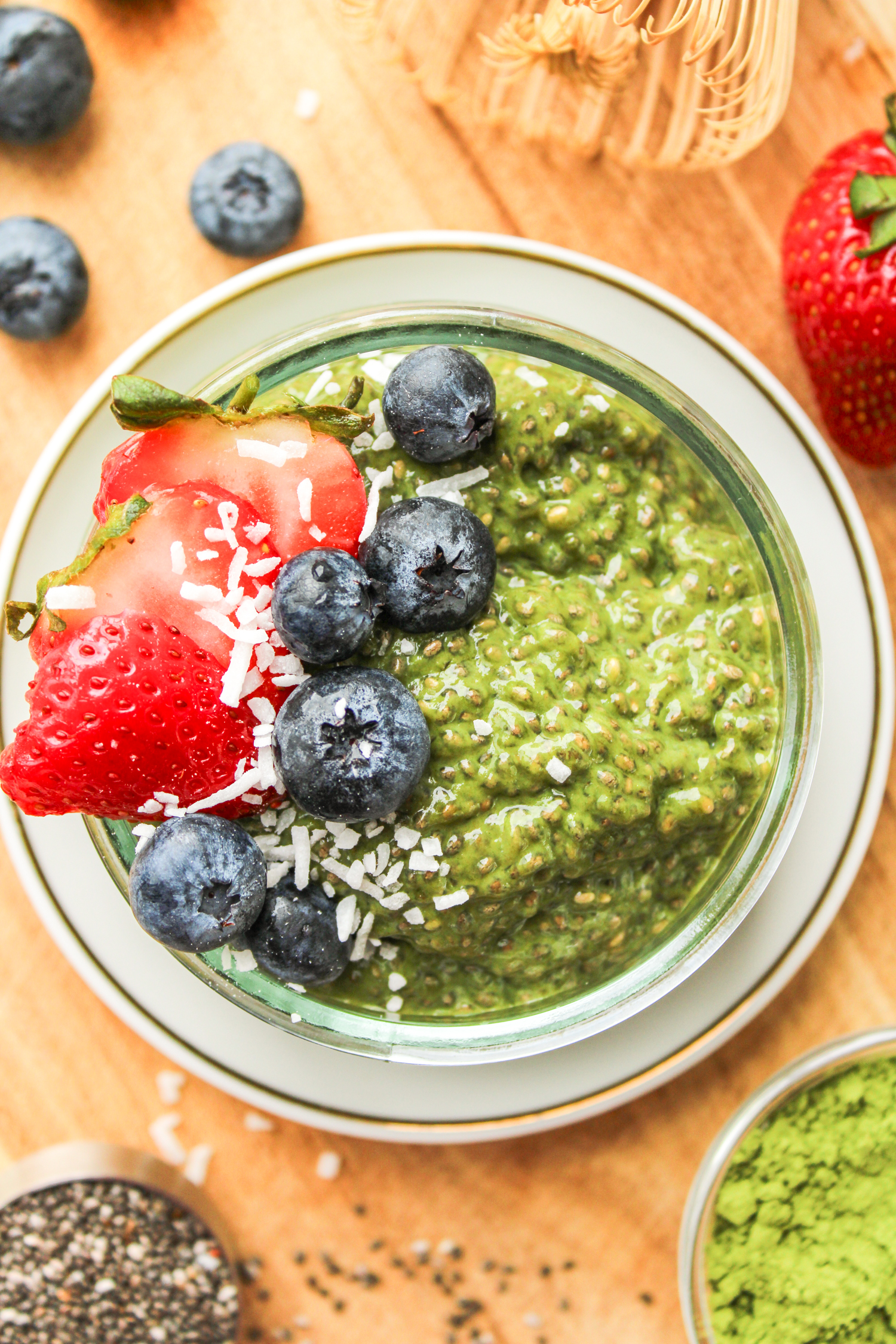 matcha chia seed pudding in a small glass bowl topped with berries and coconut flakes on top of a wooden board