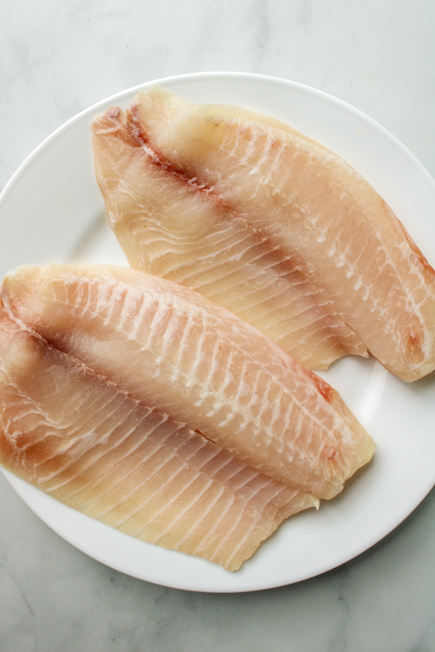 2 large fillets of fresh tilapia on a white plate prior to being breaded