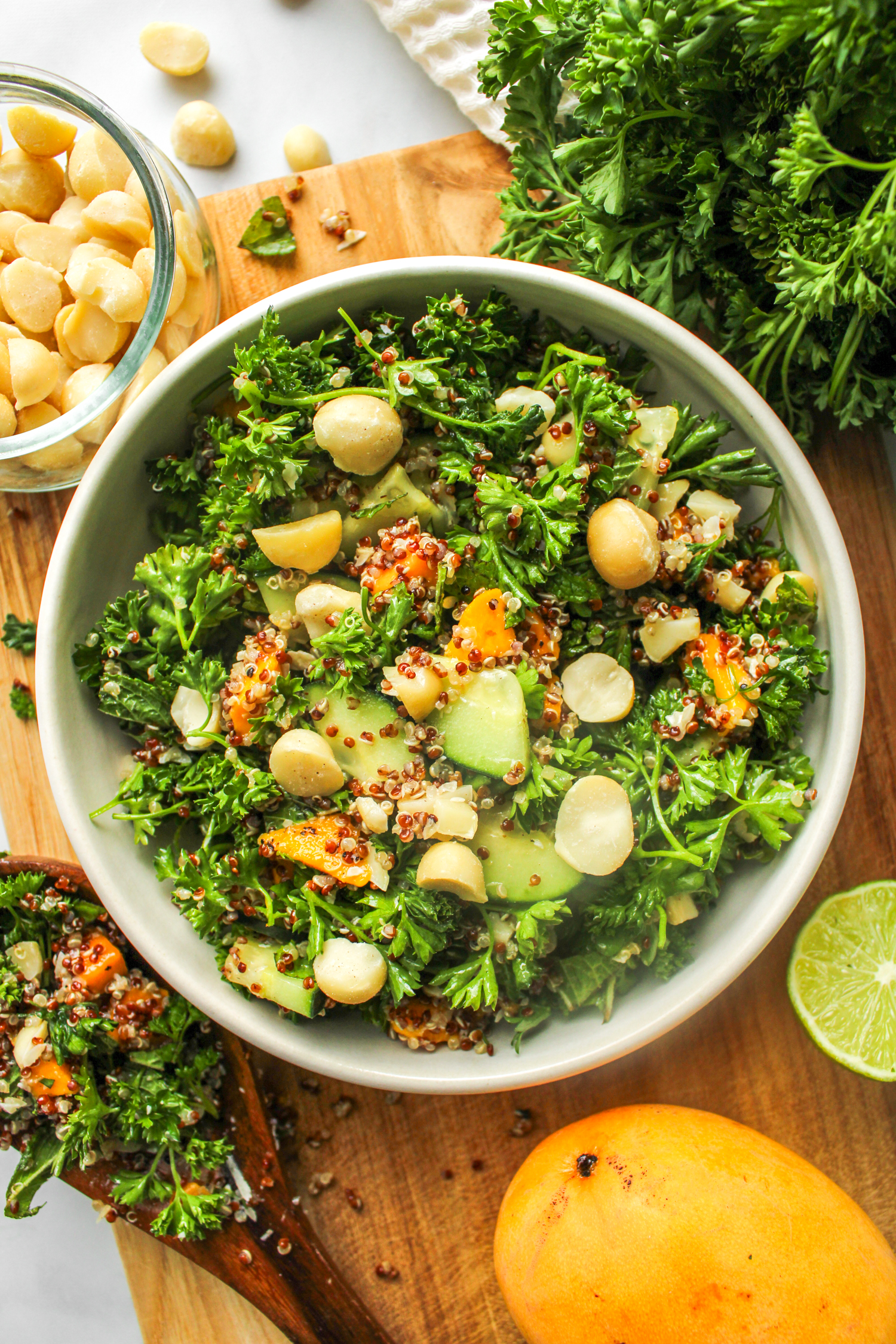 parsley quinoa salad with mango in a white ceramic bowl on top of a wooden cutting board surrounded with ingredients used