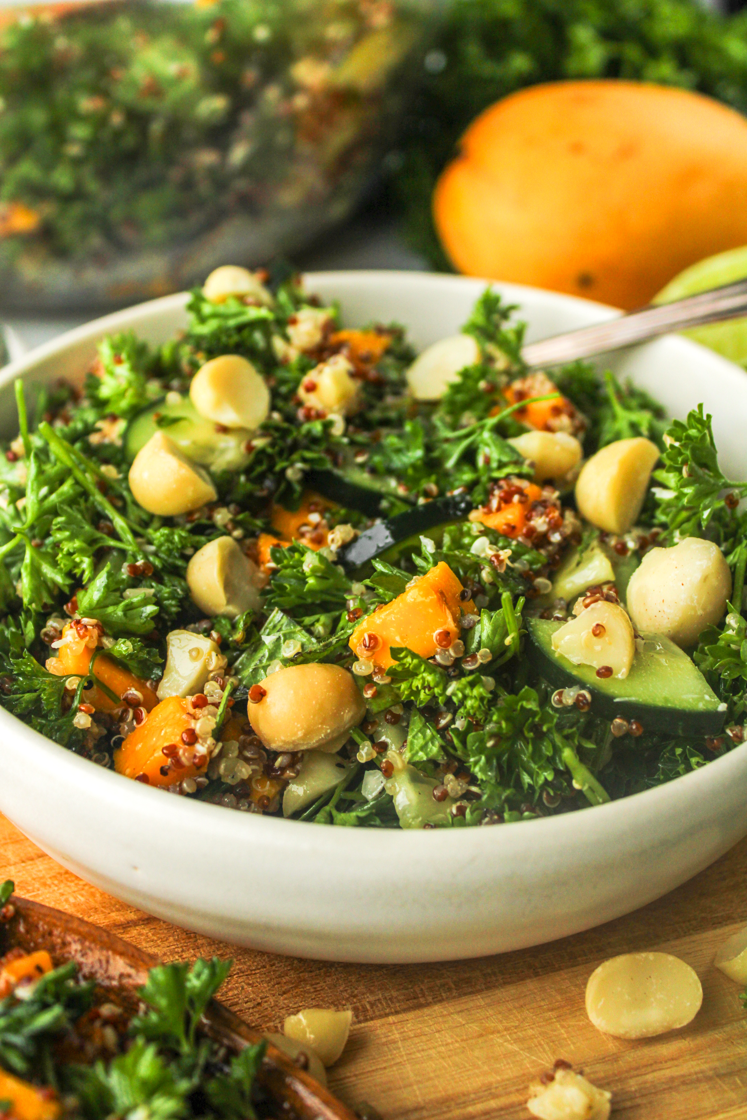 side profile parsley quinoa salad with mango in a white ceramic bowl on top of a wooden cutting board surrounded with ingredients used