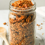 air fryer granola in a large glass mason jar on top of a white ceramic dish with a wooden spoon beside it