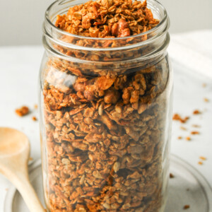 air fryer granola in a large glass mason jar on top of a white ceramic dish with a wooden spoon beside it