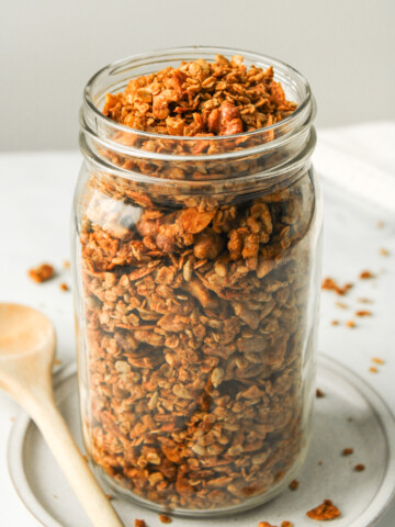 air fryer granola in a large glass mason jar on top of a white ceramic dish with a wooden spoon beside it