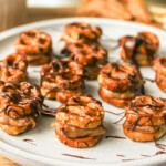angled shot of pretzel peanut butter bites drizzled with chocolate on a grey ceramic plate, surrounded by recipe ingredients
