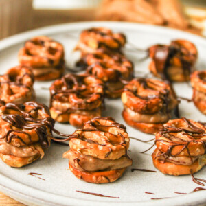 angled shot of pretzel peanut butter bites drizzled with chocolate on a grey ceramic plate, surrounded by recipe ingredients