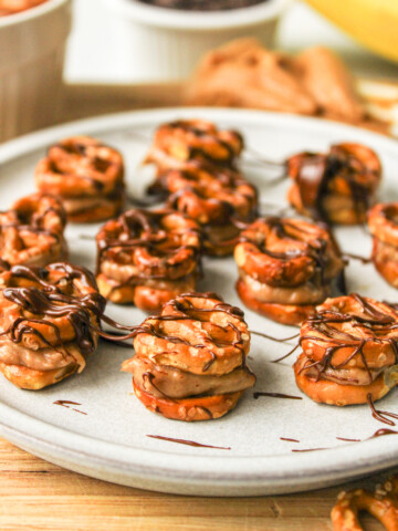angled shot of pretzel peanut butter bites drizzled with chocolate on a grey ceramic plate, surrounded by recipe ingredients