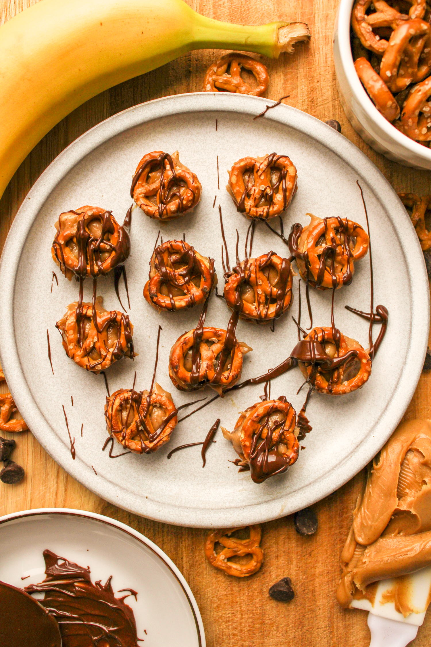 wide shot of pretzel peanut butter bites drizzled with chocolate on a grey ceramic plate, surrounded by recipe ingredients