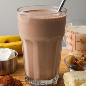 banana chocolate smoothie in a glass cup with a metal straw, surrounded by ingredients