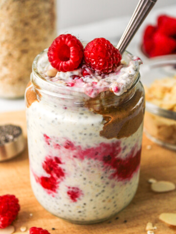 mixed overnight oats layered in a glass jar topped with raspberries, almond butter and sliced almonds