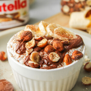 nutella overnight oats in a white ceramic dish topped with nutella, sliced banana, chopped hazelnuts and cocoa powder