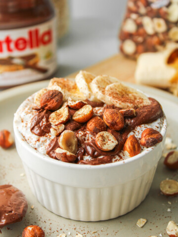 nutella overnight oats in a white ceramic dish topped with nutella, sliced banana, chopped hazelnuts and cocoa powder