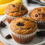 three banana chocolate chunk muffins on a grey ceramic plate with bananas and more muffins in the background