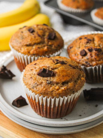 three banana chocolate chunk muffins on a grey ceramic plate with bananas and more muffins in the background