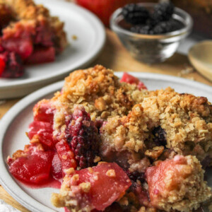 apple and blackberry crumble on a grey ceramic plate on top of a wooden cutting board with crumble and ingredients in the background