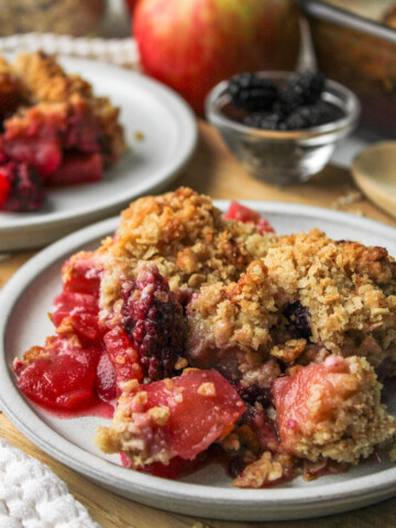 apple and blackberry crumble on a grey ceramic plate on top of a wooden cutting board with crumble and ingredients in the background