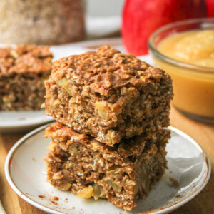 two applesauce oatmeal bars stacked on top of each other on a small white ceramic plate with ingredients in the background