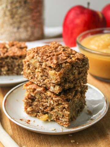 two applesauce oatmeal bars stacked on top of each other on a small white ceramic plate with ingredients in the background