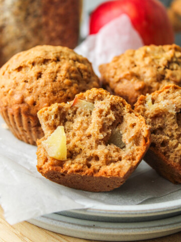 half an healthy apple oatmeal muffins on stacked white ceramic plates with other muffins surrounding it