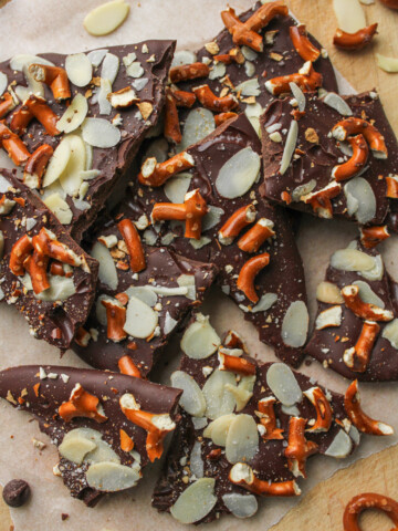 pretzel almond bark pieces on top of a wooden cutting board with pieces of pretzels, almonds and chocolate around the bark