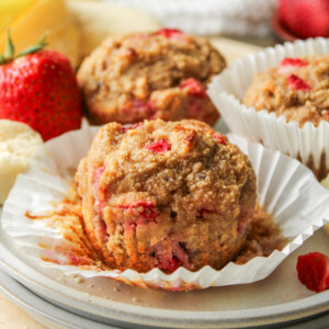 strawberry banana and almond flour muffins on a paper muffin liner on top of two grey ceramic plates with muffins in the background