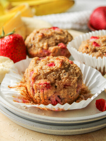 strawberry banana and almond flour muffins on a paper muffin liner on top of two grey ceramic plates with muffins in the background
