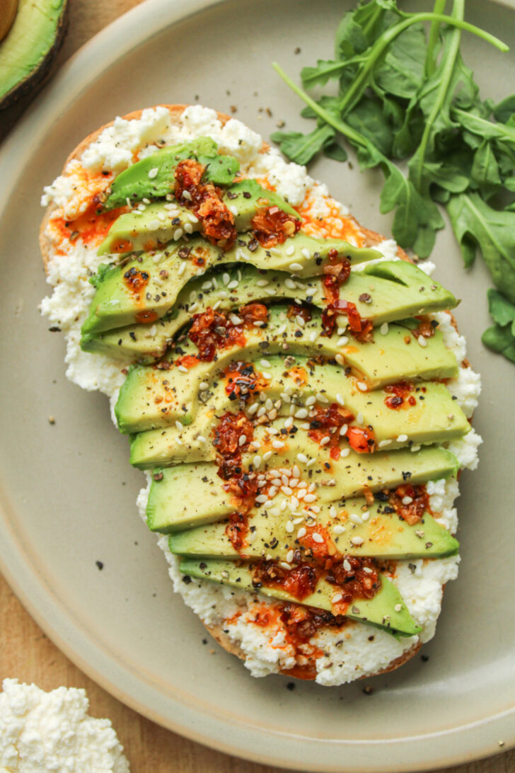 overhead shot of avocado ricotta cheese toast drizzled with chili oil on top of a ceramic plate