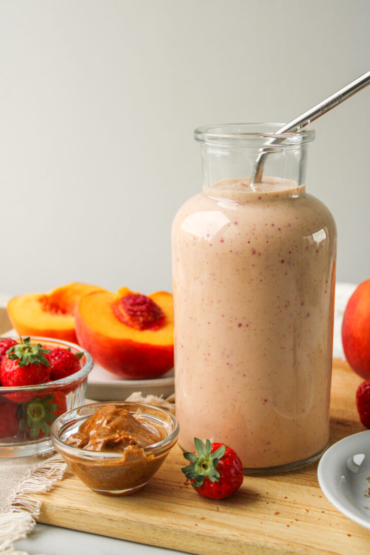 side profile of strawberry peach smoothie in a glass jug with a metal straw and ingredients on the cutting board around it