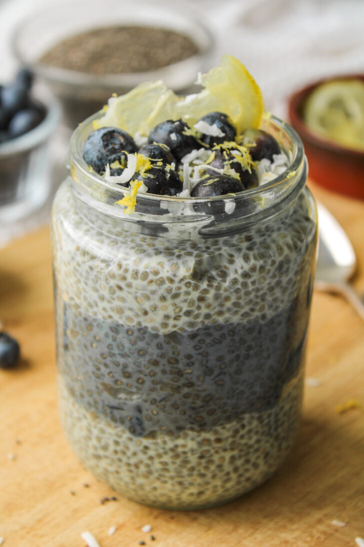 close up of lemon blueberry chia pudding layered in a glass jar, topped with blueberries, lemon zest and coconut sitting on a wooden cutting board