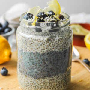 lemon blueberry chia pudding layered in a glass jar, topped with blueberries, lemon zest and coconut sitting on a wooden cutting board