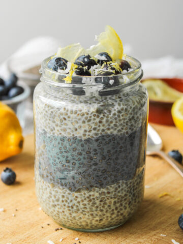 lemon blueberry chia pudding layered in a glass jar, topped with blueberries, lemon zest and coconut sitting on a wooden cutting board