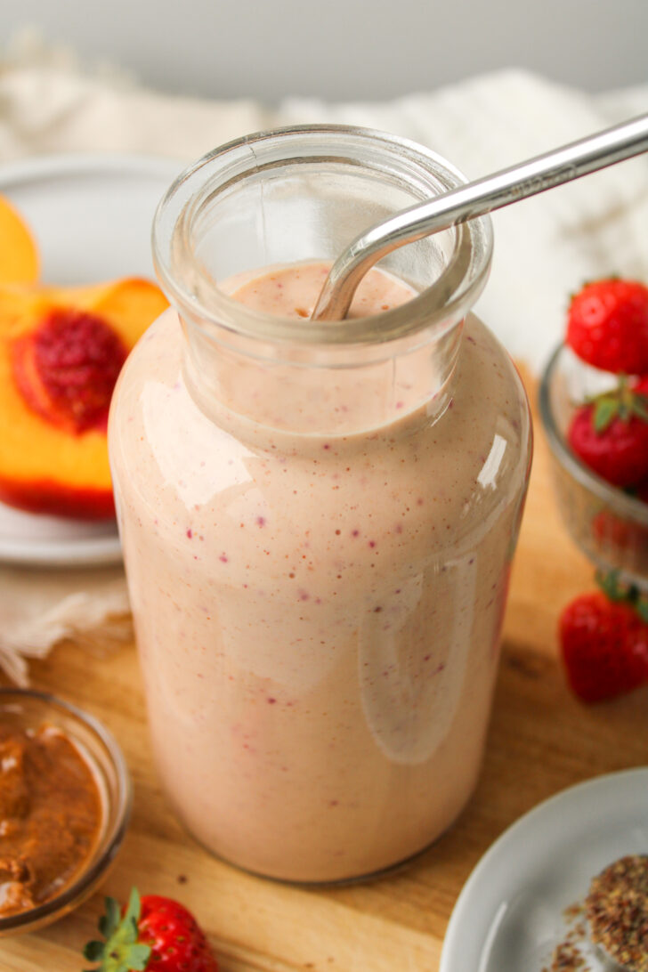 overhead angled shot of strawberry peach smoothie in a glass jug and ingredients on the cutting board around it